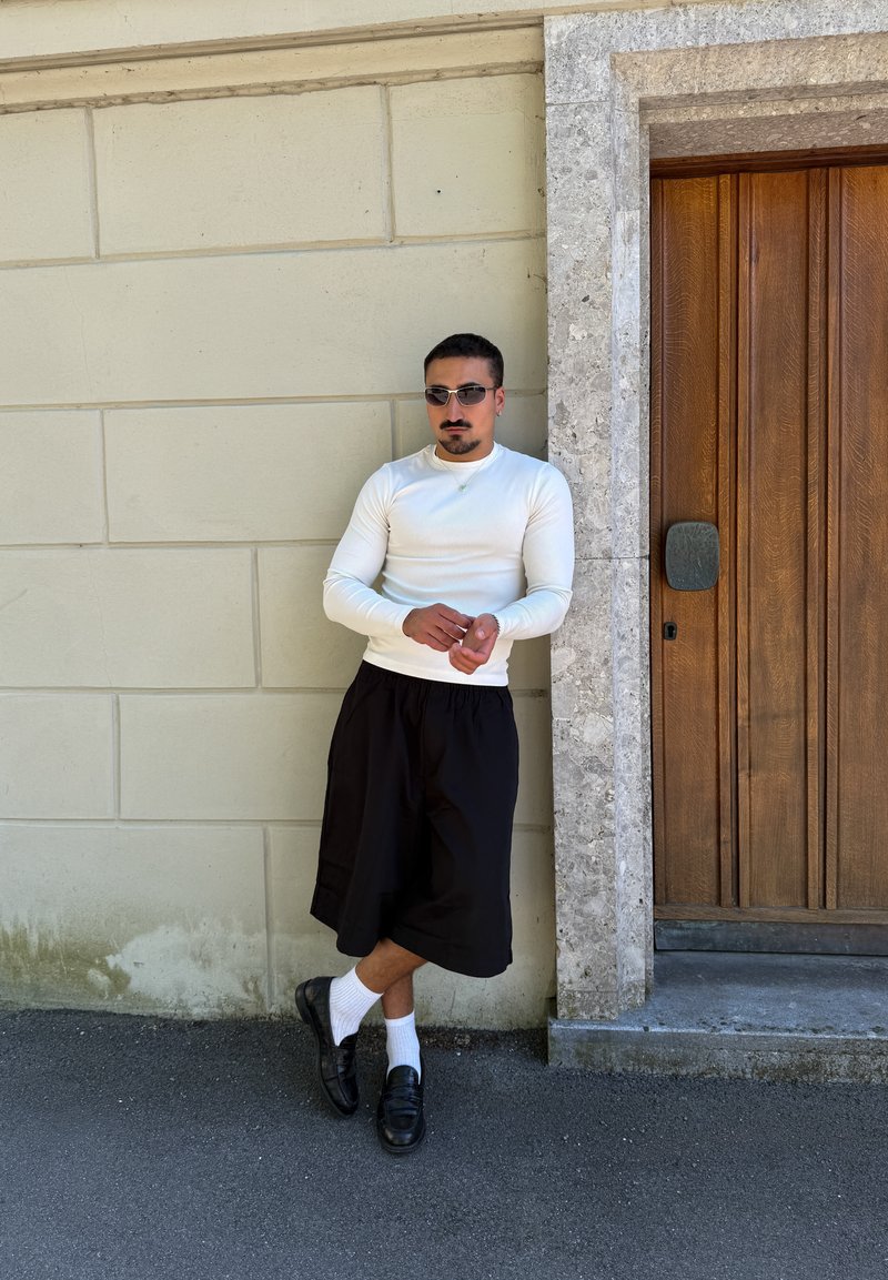 Homme portant des lunettes de soleil, appuyé contre un mur beige près d'une porte en bois, vêtu d'une chemise blanche à manches longues, d'un pantalon large noir, de chaussettes blanches et de chaussures noires.