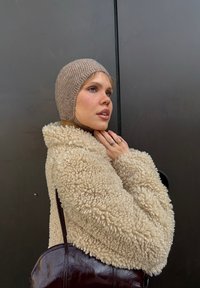 Beige, texturerad shearlingkappa med överdimensionerad krage; ljusbrun stickad mössa; mörk läderhandväska; person som poserar mot en neutral bakgrund.