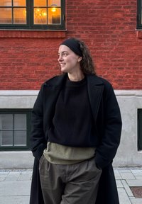 Fekete, dupla soros kabát, bordázott fekete pulóver, olívazöld, texturált derékpánt, bővebb olívazöld nadrág; vörös téglafal ablakokkal.