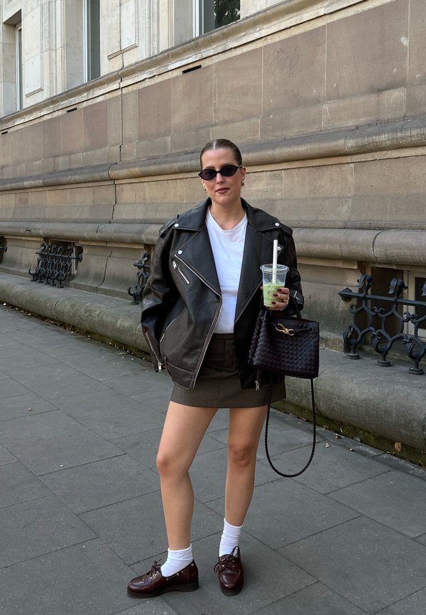 Zwarte leren motorjas, wit T-shirt, olijfgroene minirok, witte sokken en bruine veterlaarzen. Houdt een doorzichtige beker en een zwarte geweven handtas vast.