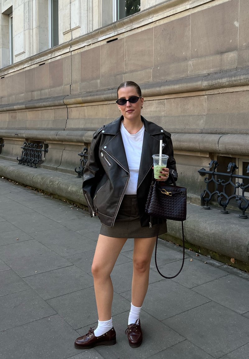 Veste de motard en cuir noir, t-shirt blanc, mini-jupe vert olive, chaussettes blanches et chaussures marron à lacets. Tenant une tasse transparente et un sac à main tissé noir.