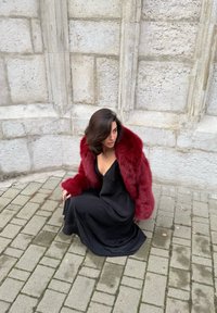 Rød imiteret pelsfrakke over en sort satin kjole, siddende på en stenbelægning, foran en tekstureret grå væg med kantede arkitektoniske detaljer.