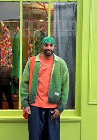 Πράσινη gradient μπλούζα πάνω από πορτοκαλί πουκάμισο, συνδυασμένη με σκούρα μπλε τζιν παντελόνια. Αξεσουάρ περιλαμβάνουν μια πράσινη μαντήλα και χρυσά κοσμήματα. Φόντο φωτεινής πράσινης βιτρίνας καταστήματος.