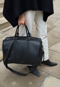 Fekete bőr duffle táska aranyszínű fém kiegészítőkkel, két felső fogantyúval és levehető vállpánttal. Simább textúrával és finom logóval rendelkezik.