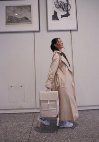 Gabardina beige con mangas largas, corte holgado y cuello ancho; mochila gris claro con solapa, bolsillos frontales dobles y correas ajustables.