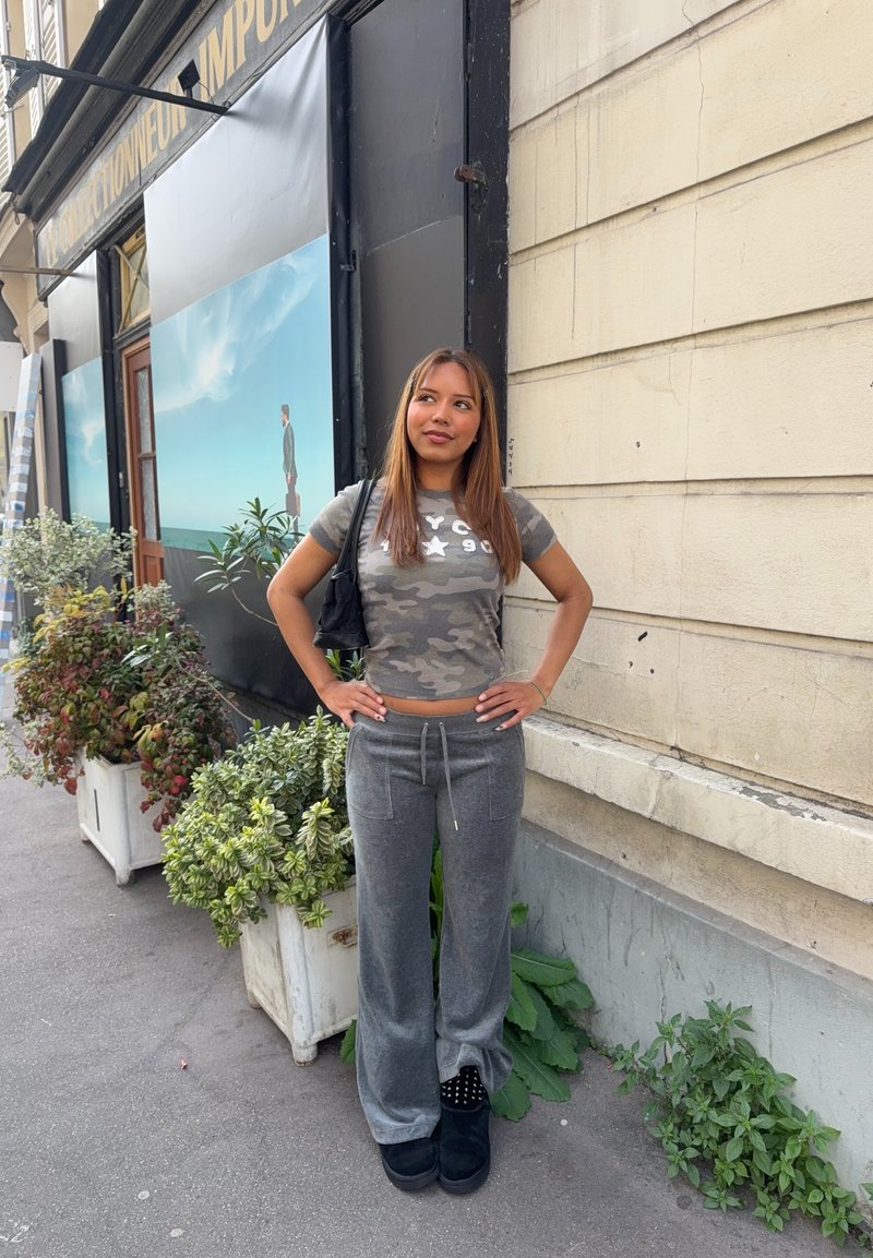 Νεαρή γυναίκα με μακριά μαλλιά, με γκρι τοπ και παντελόνι παραλλαγής, στέκεται με τα χέρια στη μέση σε πεζοδρόμιο της πόλης κοντά σε γλαστράκια και τοίχο κτιρίου.