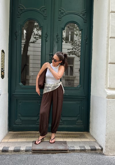 Mujer con gafas de sol de pie junto a una puerta verde ornamentada, vestida con camiseta de tirantes blanca, pantalones marrones, sandalias y lleva un bolso marrón al hombro.