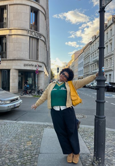 Suéter verde con letras blancas debajo de una chaqueta bomber beige, pantalones anchos oscuros y bolsa de hombro naranja. Calle de adoquines con edificios.