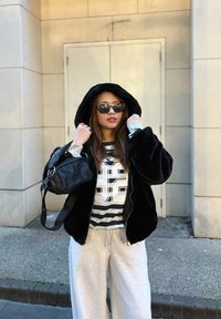 Chaqueta con capucha de piel negra, camiseta a rayas en gris y blanco con gráfico, pantalones deportivos grises y mochila negra. Las gafas de sol completan el look.