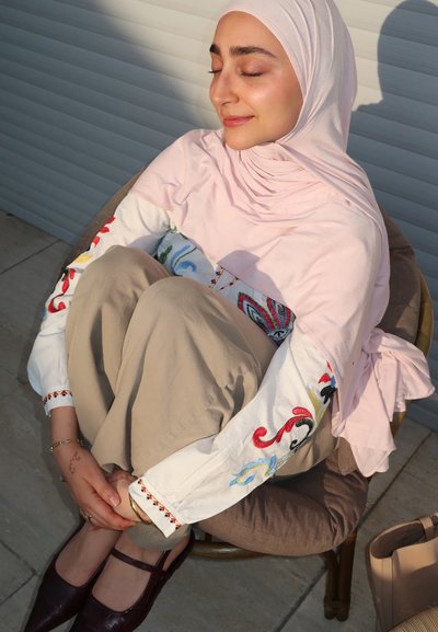 Mujer con hiyab rosa y blusa blanca bordada sentada con las piernas cruzadas en una silla, con los ojos cerrados y sonriendo suavemente a la luz del sol.