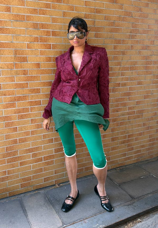 Personne portant de grandes lunettes de soleil réfléchissantes, un blazer bordeaux texturé, une tunique verte, des leggings verts courts et des chaussures plates noires, debout contre un mur en briques.