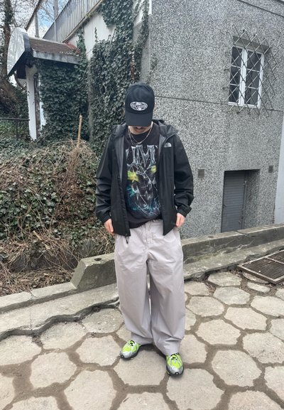 Persona con gorra negra, camiseta negra con gráficos, chaqueta negra, pantalones anchos gris claro y zapatillas verde neón de pie sobre un pavimento de piedra cerca de una pared cubierta de hiedra.