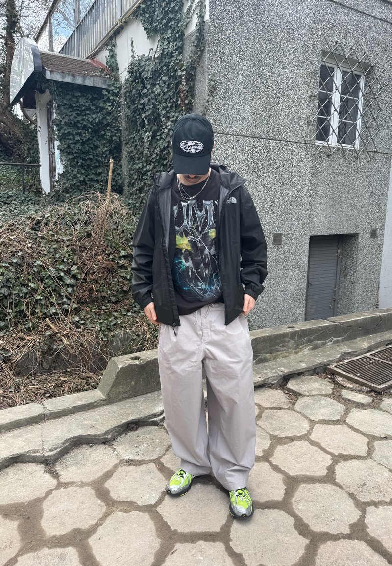 Personne portant une casquette noire, un t-shirt graphique noir, une veste noire, un pantalon large gris clair et des baskets vert fluo, debout sur un pavement en pierre près d'un mur couvert de lierre.