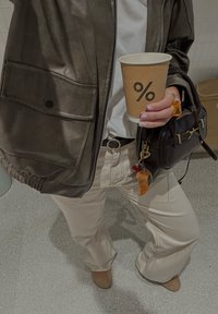 Hnědá bunda z umělé kůže, bílé tričko, krémové široké kalhoty a béžové kotníkové boty. Drží papírový kelímek na kávu s kulatým logem.