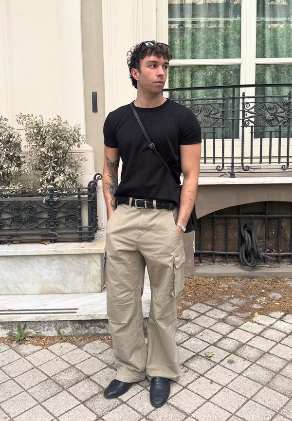 Junger Mann mit tätowierten Armen, der ein schwarzes T-Shirt, beige Cargohosen und schwarze Schuhe trägt, steht auf einem gefliesten Gehsteig in der Nähe eines Gebäudes mit Fenstern.