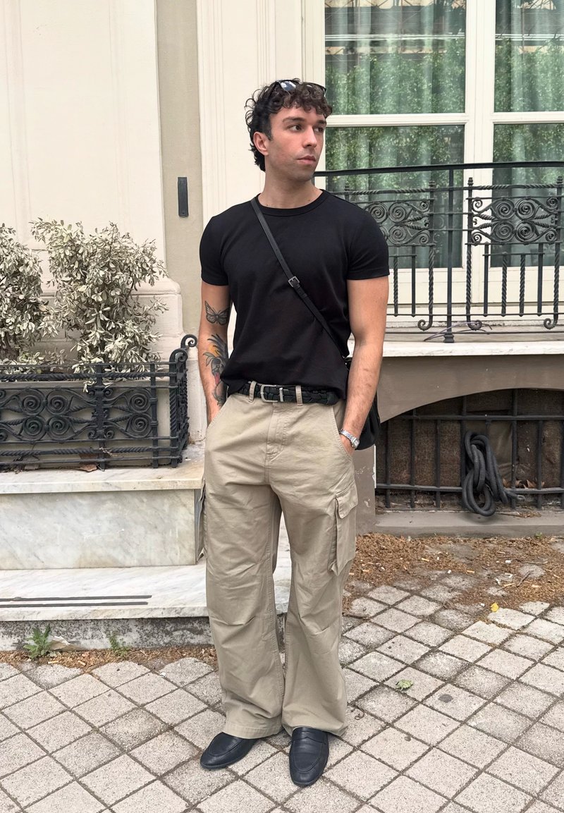 Jeune homme aux bras tatoués portant un t-shirt noir, un pantalon cargo beige et des chaussures noires, debout sur un trottoir carrelé près d'un bâtiment vitré.