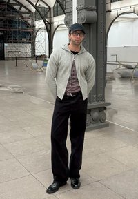 Ljusgrå zip-up cardigan över en mörk t-shirt, svarta vida jeans och svarta skor. Placerad i en rymlig industriell miljö med byggställningar.