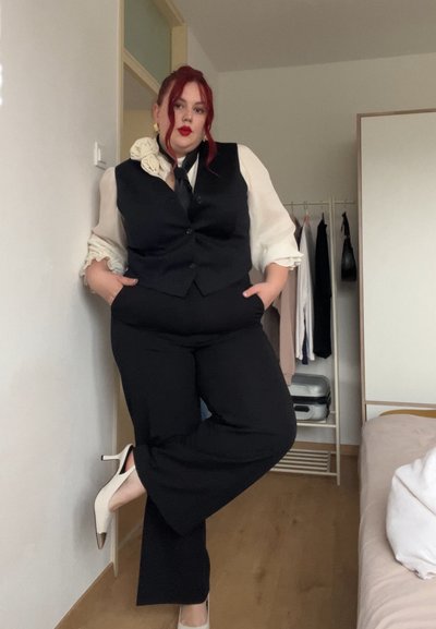 Chaleco negro ajustado y pantalones de pierna ancha combinados con una blusa blanca con puños con volantes y un adorno de flor de tela. Tacones blancos con puntas doradas.