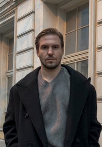 Juodas vilnonis paltas, po juo pilkas korėtas megztinis, stovintis prie šviesios marmuro sienos. Yra su skeltu apykakle ir mygtukų akcentais.