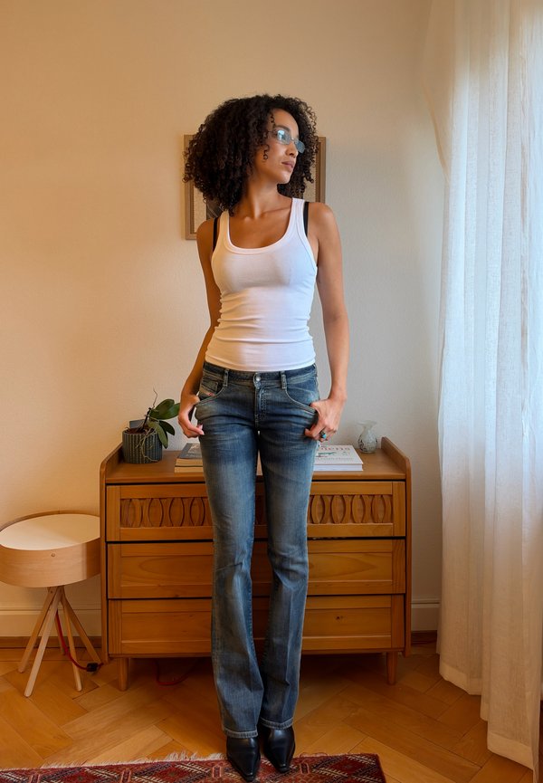 Une femme portant un débardeur blanc, un jean bleu et des lunettes se tient près d’une commode en bois et regarde vers une fenêtre aux rideaux tirés.