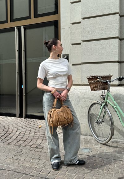 Conjunto informal compuesto por una camiseta blanca ajustada y jeans sueltos de color azul claro. Bolso marrón texturado con detalles de flecos. Bicicleta verde cercana.