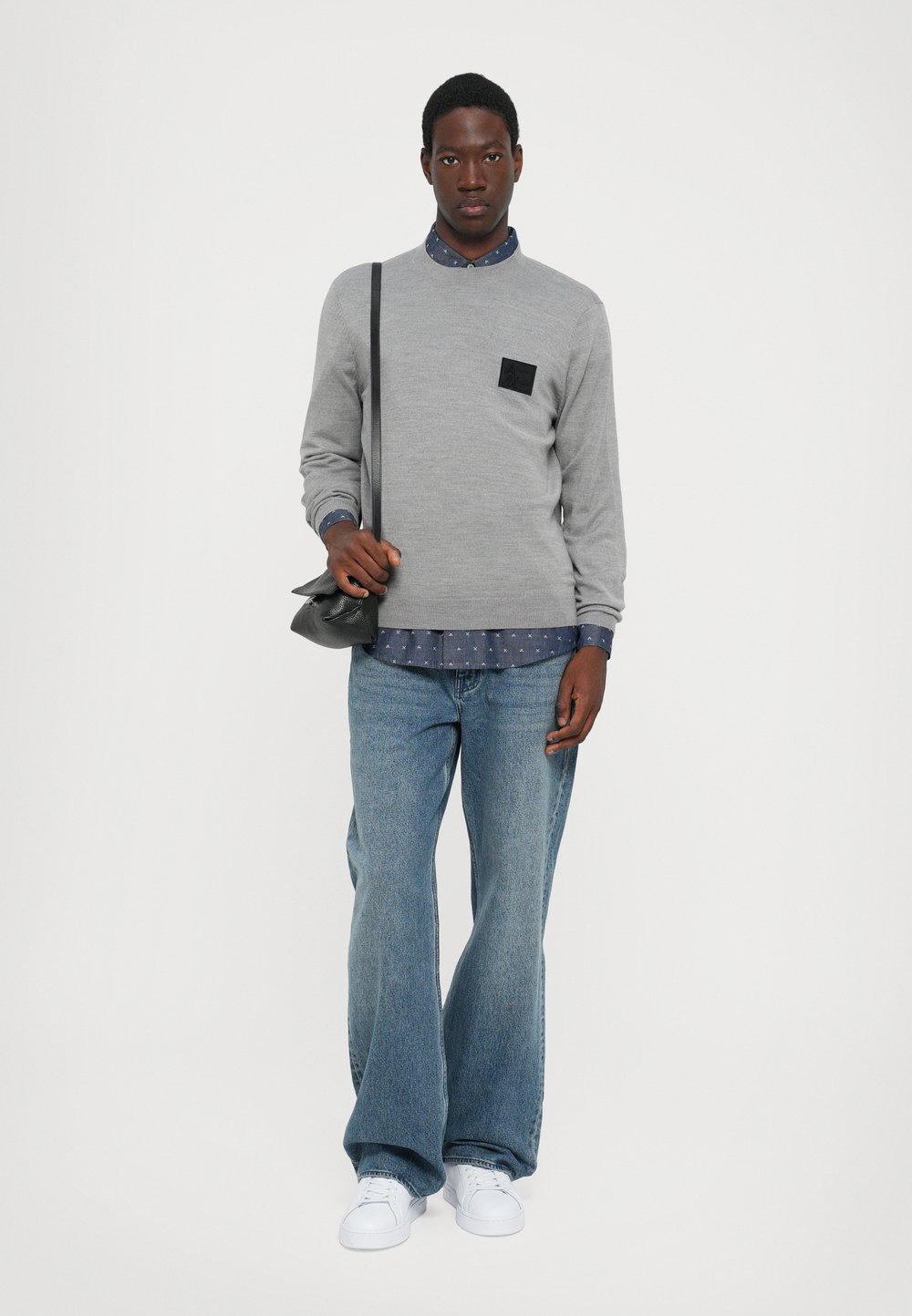 Maglione grigio con una toppa nera, indossato sopra una camicia blu a pois, abbinato a jeans ampi in denim e sneakers bianche. Borsa a tracolla nera.