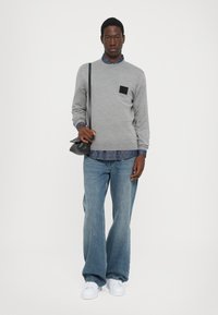 Maglione grigio con una toppa nera, indossato sopra una camicia blu a pois, abbinato a jeans ampi in denim e sneakers bianche. Borsa a tracolla nera.
