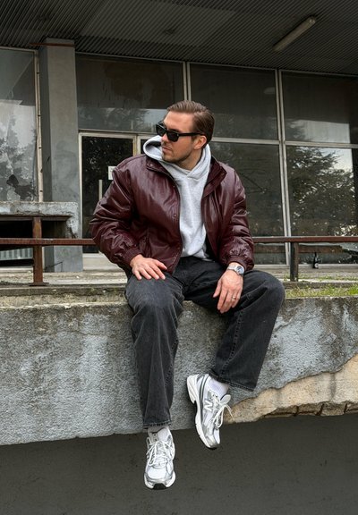 Hombre con gafas de sol, chaqueta de cuero burdeos, sudadera con capucha gris, pantalones negros y zapatillas blancas sentado en un bordillo de hormigón al aire libre.
