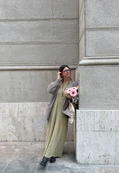 Mujer con gafas y un vestido largo con chaqueta, sosteniendo flores rosas, apoyada contra una pared de piedra gris al aire libre.