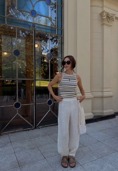 Top sin mangas a rayas en blanco y negro, combinado con pantalones de lino blancos holgados. Los accesorios incluyen un bolso de mimbre y sandalias marrones.