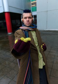 Abrigo marrón a cuadros con bufanda multicolor vibrante, guantes morados y detalles con acentos amarillos. Ambiente urbano con pavimento mojado.