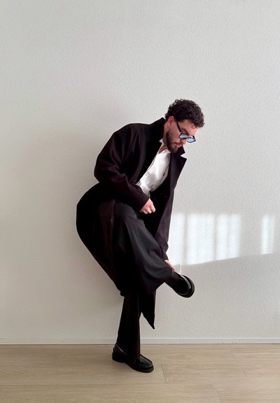 Hombre con abrigo negro, camisa blanca y zapatos negros, ajustándose el zapato mientras está de pie contra una pared blanca con reflejos de luz.