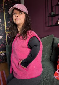 Femme aux longs cheveux noirs portant une casquette rose et un gilet polaire rose, assise de côté sur un canapé vert dans une pièce sombre à fleurs et violette.