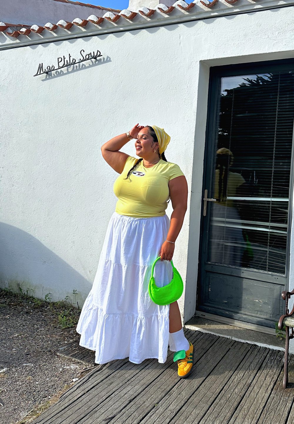 Κίτρινο εφαρμοστό μπλουζάκι με λογότυπο, λευκή tiered μακριά φούστα, πράσινη τσάντα και πολυχρωμα sneakers, μπροστά από έναν λευκό τοίχο και πόρτα.