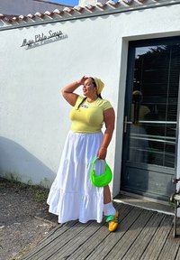 Κίτρινο εφαρμοστό μπλουζάκι με λογότυπο, λευκή tiered μακριά φούστα, πράσινη τσάντα και πολυχρωμα sneakers, μπροστά από έναν λευκό τοίχο και πόρτα.