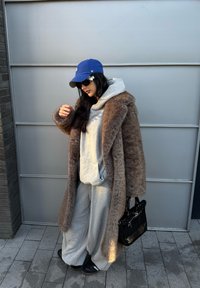 Cappotto in pelliccia sintetica marrone su una felpa grigia e pantaloni della tuta. Cappello blu, occhiali da sole neri e una piccola borsa nera. Ambientazione urbana con pavimentazione.