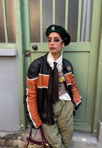 Musta nahkatakki oransseilla yksityiskohdilla, valkoinen paita, pilkullinen solmio, oliivinvihreät housut, vihreä baretti ja kirkkaat lasit, seisoo vihreän oven edessä.