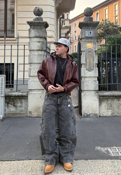 Chaqueta de cuero marrón con cremallera, camiseta negra, pantalones de mezclilla grises anchos con bolsillos y botas marrones; de pie al aire libre cerca de una puerta de piedra.