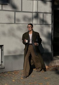 Γυναίκα ντυμένη με ένα φαρδύ σκούρο παλτό και καφέ παντελόνια με φαρδιά γραμμή, περπατάει στο πεζοδρόμιο κρατώντας ένα φλιτζάνι καφέ και μια μαύρη τσάντα, κάτω από τον ήλιο.