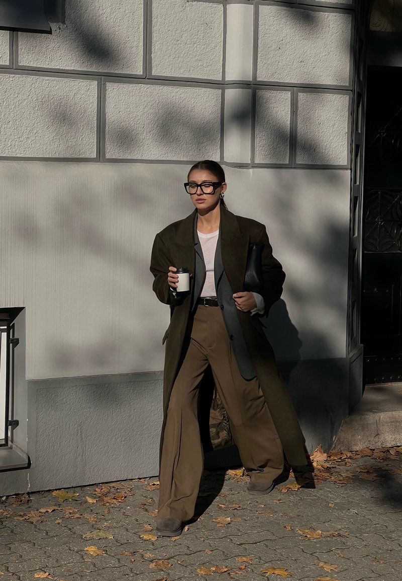 Γυναίκα ντυμένη με ένα φαρδύ σκούρο παλτό και καφέ παντελόνια με φαρδιά γραμμή, περπατάει στο πεζοδρόμιο κρατώντας ένα φλιτζάνι καφέ και μια μαύρη τσάντα, κάτω από τον ήλιο.