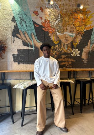 Modelo luciendo una camisa blanca de manga larga y pantalones beige de pierna ancha, sosteniendo gafas de sol. El fondo presenta un mural colorido y taburetes negros.