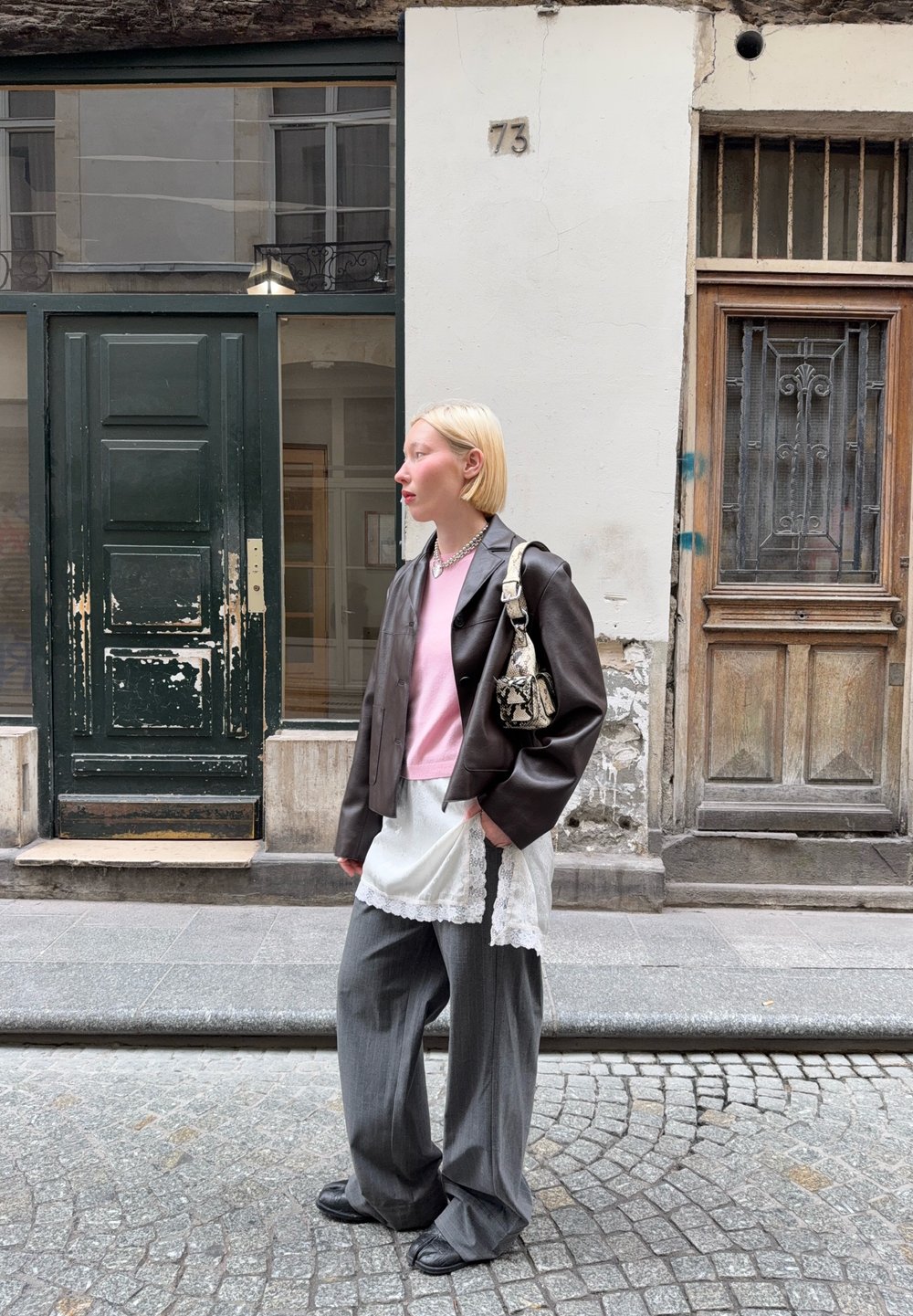 Brown leather cropped jacket, pink jumper, white lace-trimmed skirt, grey wide-leg trousers, black shoes, patterned handbag. Urban backdrop.