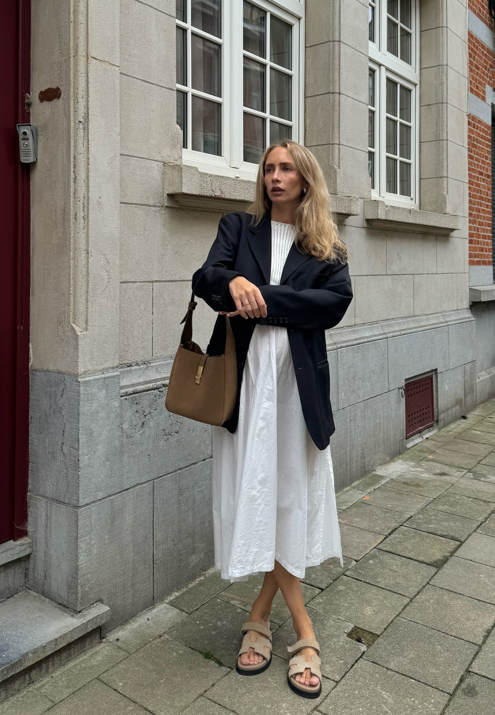 Blazer oversized preto, top de malha canelada branca, vestido longo branco, mala de ombro castanha e sandálias bege com solas pretas, em pé sobre uma calçada de pedras.