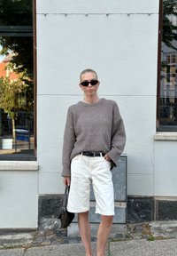 Καφέ πλεκτό πουλόβερ με φαρδιές μανίκες, κρεμ σορτς, μαύρη ζώνη και ηλιόφωνα. Κρατώντας μια μικρή μαύρη τσάντα. Στεκόμενη μπροστά σε ένα ανοιχτό τοίχο.