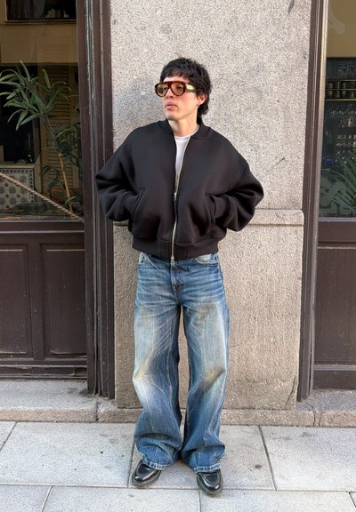 Persona que lleva una chaqueta negra con cremallera extragrande, vaqueros azules holgados, zapatos negros y grandes gafas de sol de pie junto a una pared de piedra gris.