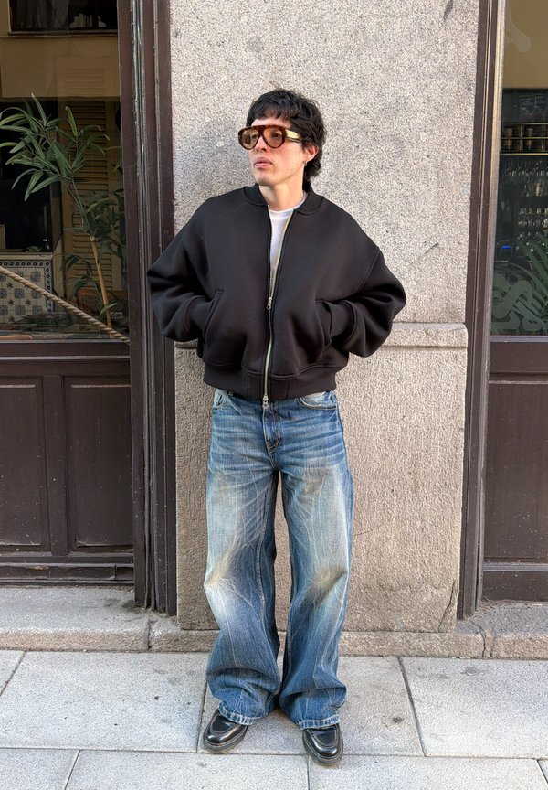 Person iført en oversized sort lynlåsjakke, løse blå jeans, sorte sko og store solbriller, der står op ad en grå stenvæg.