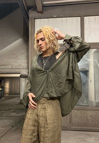 Olive zelena oversized majica s dugim rukavima, mekana, lepršava tkanina; nosi se preko crnog topa bez rukava i uparena je s svijetlosmeškim, nabranim hlačama.