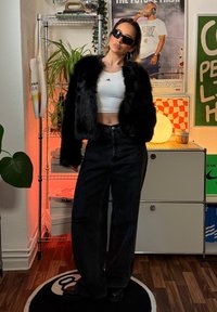 Jachetă neagră din blană sintetică peste un top alb scurt, asortată cu blugi negri cu talie înaltă și ghete negre masive; cadru interior cu plante și decorațiuni.