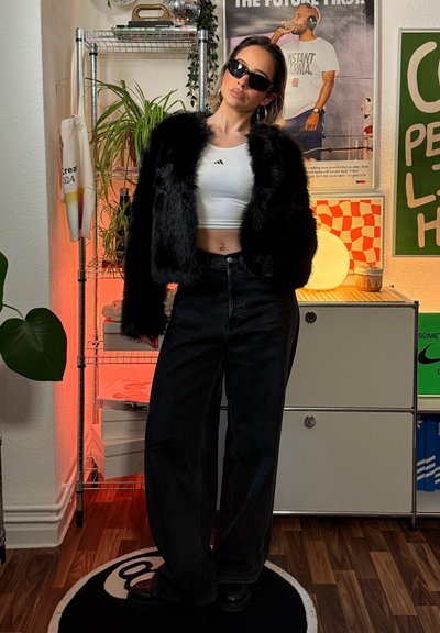 Chaqueta de piel sintética negra sobre un top corto blanco, combinada con vaqueros negros de talle alto y botas negras gruesas; ambiente interior con plantas y decoración.