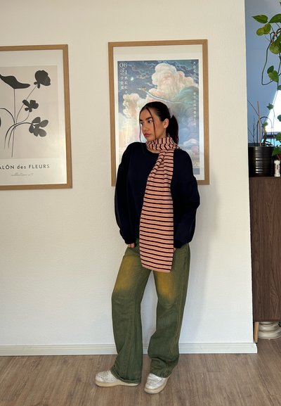 Joven con suéter negro holgado, pantalones verdes, zapatillas blancas y bufanda rosa a rayas, de pie en el interior contra una pared con obras de arte enmarcadas.